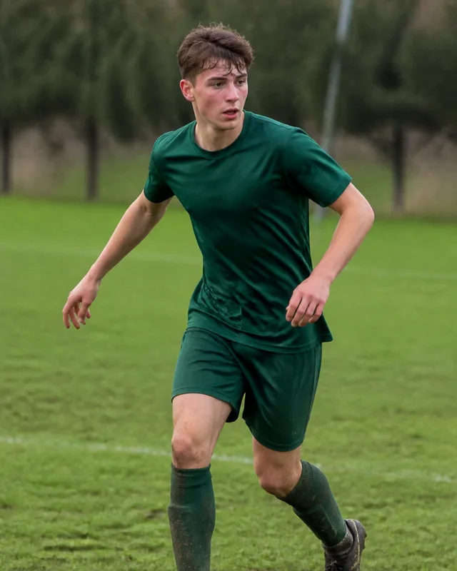 A teenage boy running with a football on a grass pitch